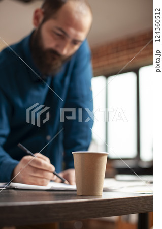 Closeup of brown paper cup placed on table as male analyst holding pencil and reading company documents. Selective focus on coffee mug on office desk as businessman reviews financial paperwork nearby. Closeup of brown paper cup placed on table as male analyst holding pencil and reading company documents. Selective focus on coffee mug on office desk as businessman reviews financial paperwork nearby. 124360512