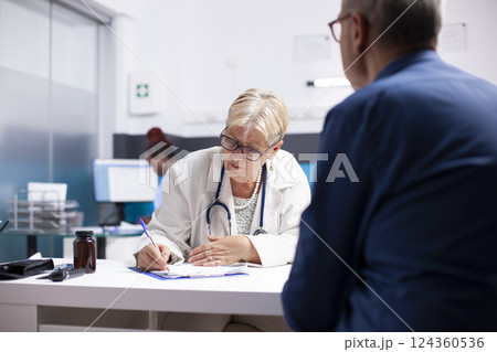 Senior medic specialist using clipboard to write prescription for elderly man at checkup. Retired female doctor taking notes during medical consultation with male pensioner patient in hospital office. 124360536