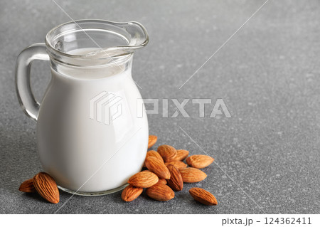 Fresh almond milk in glass pitcher stand on gray background with nuts scattered around it, creating visually appealing scene that promotes healthy eating and vegan alternatives Fresh almond milk in glass pitcher stand on gray background with nuts scattered around it, creating visually appealing scene that promotes healthy eating and vegan alternatives 124362411