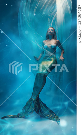 Mysterious elegant young woman, mermaid with golden tail, covered in flying fabric, floating against dreamlike ocean background 124364587