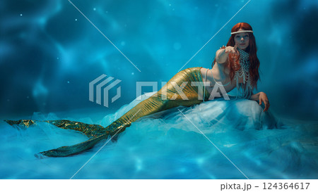 Enticing gesture. Redhaired mermaid with shimmering golden tail floating in soft blue light, exuding elegance and mystery 124364617