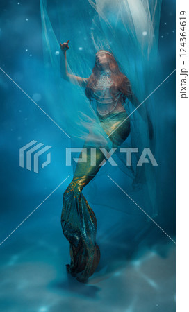 Flying towards dreams. Graceful young woman, redhaired mermaid elegantly covered in transparent veil, dancing against blue ocean-like background. 124364619