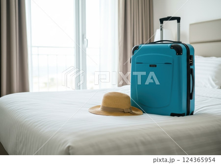 Bright blue suitcase and straw hat on hotel bed by sunny window 124365954