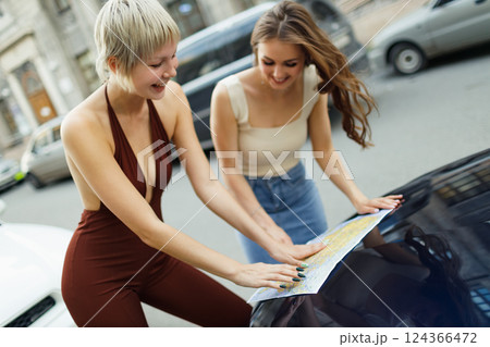 Happy young two women tourist travelers with paper map at rental car vehicle bonnet is exploring new city Happy young two women tourist travelers with paper map at rental car vehicle bonnet is exploring new city 124366472