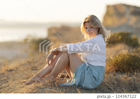 Blonde woman relaxing on a hill at sunset by the sea Blonde woman relaxing on a hill at sunset by the sea 124367122