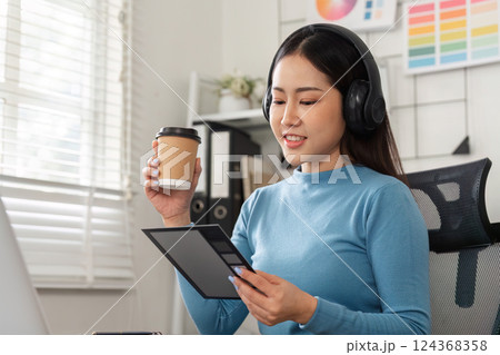 Design Review and Coffee Enjoyment. A woman in headphones examining design concepts while sipping coffee, illustrating a modern creative routine. 124368358
