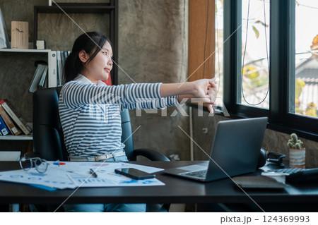 A young woman stretches at her office desk, surrounded by a laptop and documents, in a modern workspace with natural light. 124369993