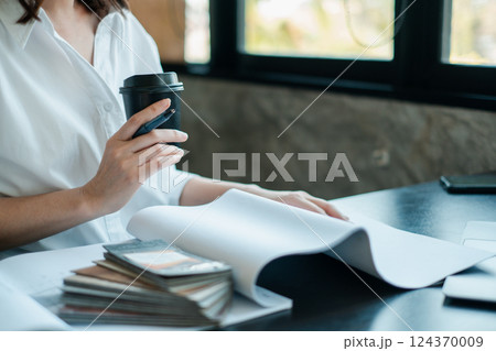 Professional woman in a white shirt holding a coffee cup while reviewing documents at a modern office desk. Professional woman in a white shirt holding a coffee cup while reviewing documents at a modern office desk. 124370009