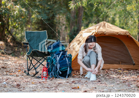 Woman tying shoes near a tent in a forest setting, surrounded by camping gear and nature. Woman tying shoes near a tent in a forest setting, surrounded by camping gear and nature. 124370020