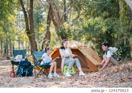 A family camping in the forest, sitting by a tent, enjoying nature and outdoor activities together. A family camping in the forest, sitting by a tent, enjoying nature and outdoor activities together. 124370023