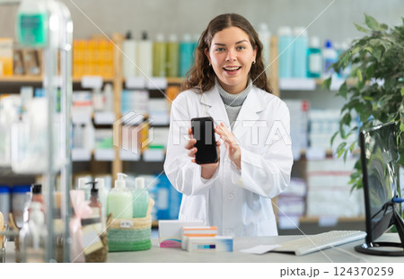Smiling female pharmacist showing screen of her smartphone. Offers to install application on your smartphone 124370259