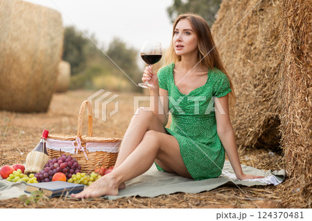 Young beautiful woman in summer green dress sitting and drinking on soft mat near stack of straw with glass of wine, romantic picnic in field Young beautiful woman in summer green dress sitting and drinking on soft mat near stack of straw with glass of wine, romantic picnic in field 124370481