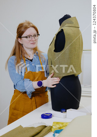 female fashion designer seamstress with red hair tries on clothes for a mannequin. female fashion designer seamstress with red hair tries on clothes for a mannequin. 124370804