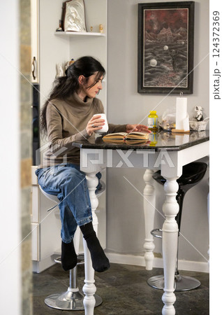 A woman is savoring a warm beverage as she immerses herself in the pages of a captivating book, surrounded by the cozy ambiance of her inviting kitchen. A woman is savoring a warm beverage as she immerses herself in the pages of a captivating book, surrounded by the cozy ambiance of her inviting kitchen. 124372369