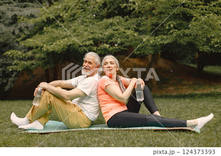 Senior man and woman with yoga mats sitting at park with bottles of water. Positive mature man and his elderly wife after fitness or yoga class in nature. Wellness and healthy lifestyle on retirement. 124373393