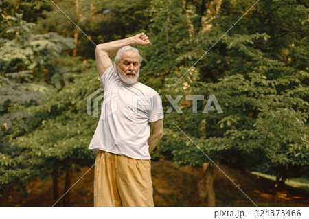 Senior man in white t-shirt standing at park and doing excercises. Positive mature woman has fitness or yoga class in nature. Wellness and healthy lifestyle on retirement. 124373466