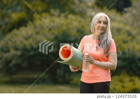 Senior woman with yoga mat standing at park with bottle of water. Positive mature woman after fitness or yoga class in nature. Wellness and healthy lifestyle on retirement. Senior woman with yoga mat standing at park with bottle of water. Positive mature woman after fitness or yoga class in nature. Wellness and healthy lifestyle on retirement. 124373520