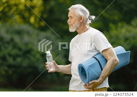 Senior man with yoga mat standing at park with bottle of water. Positive mature man after fitness or yoga class in nature. Wellness and healthy lifestyle on retirement. Senior man with yoga mat standing at park with bottle of water. Positive mature man after fitness or yoga class in nature. Wellness and healthy lifestyle on retirement. 124373524