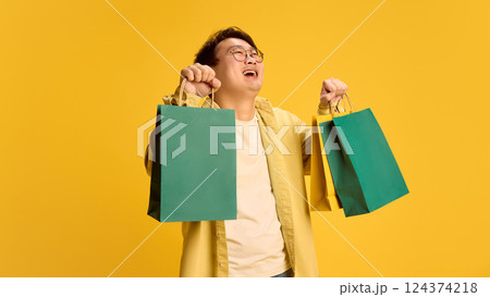 Portrait of happy, cheerful young Asian guy, dressed casual attire, and holding shopping bags with joyful expression against sunny yellow background. 124374218