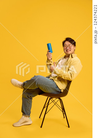 Happy young Asian man sitting on chair and showing thumb up and can with empty space against sunny yellow background. Happy young Asian man sitting on chair and showing thumb up and can with empty space against sunny yellow background. 124374221