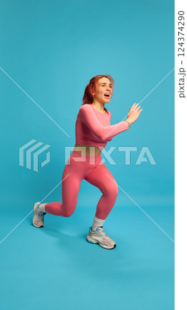 Smiling woman in pink activewear doing jumping lunge with powerful movement, embodying joy and effort in sport. 124374290