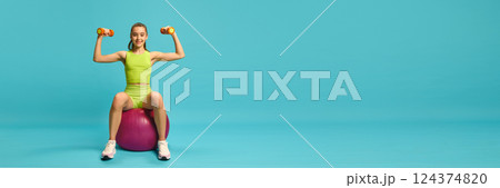 Banner. Charming, athletic girl, sitting on fitness ball and doing exercises for hands with dumbbells against blue background with copy space. Banner. Charming, athletic girl, sitting on fitness ball and doing exercises for hands with dumbbells against blue background with copy space. 124374820