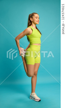 Full length photo of young girl, doing stretching exercises for legs, balancing on one leg against bright blue studio background. Full length photo of young girl, doing stretching exercises for legs, balancing on one leg against bright blue studio background. 124374821