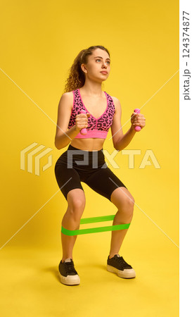 Young woman doing resistance band squat with dumbbells, training legs and arms simultaneously in energetic workout scene. 124374877