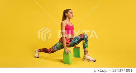 Confident young girl in lunge pose with yoga blocks, improving balance and hip mobility in bold activewear against yellow background. Confident young girl in lunge pose with yoga blocks, improving balance and hip mobility in bold activewear against yellow background. 124374891