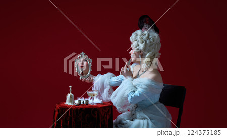 Mirror scene. Medieval noblewoman painting lips with lipstick, looking into decorated mirror, surrounded by vanity items 124375185