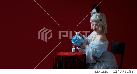Playful ritual. Rococo-styled young woman pouring milk from blue jug into glass against deep red background. Playful ritual. Rococo-styled young woman pouring milk from blue jug into glass against deep red background. 124375188