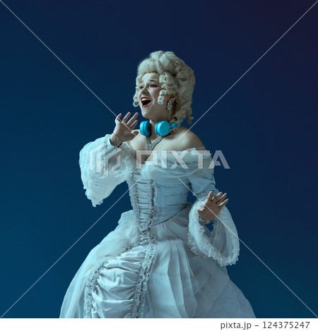 Elegant young woman in image of medieval duchess, dressed in white dress and wig, with modern headphones singing on blue background. 124375247