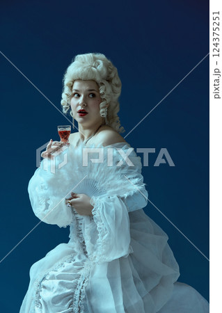 Surprised moment. Elegant woman holding glass with alcohol and fan, eyes wide in dramatic expression. 124375251