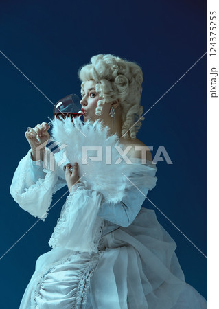 Hidden glances. Baroque woman sipping red wine behind feather fan against blue background. 124375255
