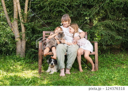 Grandmother sitting on a wooden bench hugging granddaughters with pet cat and dog in a summer garden Grandmother sitting on a wooden bench hugging granddaughters with pet cat and dog in a summer garden 124376318