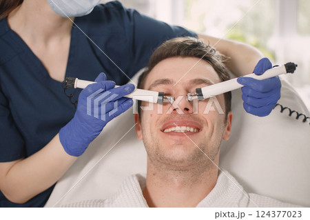 Close up of a young man laying in cosmetologist cabinet and has a cavitation procedure for face skin. Female cosmetologist wearing blue medical costume and gloves. Man wearing white bathrobe. Close up of a young man laying in cosmetologist cabinet and has a cavitation procedure for face skin. Female cosmetologist wearing blue medical costume and gloves. Man wearing white bathrobe. 124377023