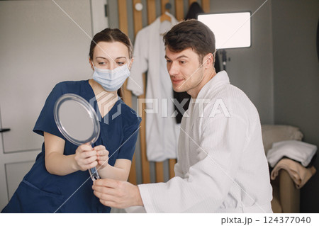 Man sitting in cosmetologist cabinet and looking in the mirror. Female cosmetologist wearing blue medical costume and face mask. Man wearing white bathrobe. 124377040
