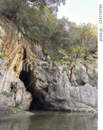 Rocky cave entrance surrounded by lush greenery and calm water reflecting nature's beauty concept 124379798