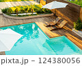 Elegant turquoise swimming pool with wooden deck, lounge chairs, and umbrellas in a sunny backyard 124380056