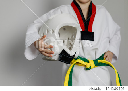 A child in a taekwondo uniform holds in his hands a white protective helmet for his head, ready for classes. Yellow-green belt A child in a taekwondo uniform holds in his hands a white protective helmet for his head, ready for classes. Yellow-green belt 124380391