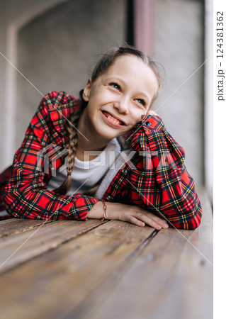 Vertical portrait of cheerful preteen girl with braided hair, smiling brightly looking away. Pretty 10 years old girl wearing red checkered shirt leaning against table by window, natural day light. 124381362