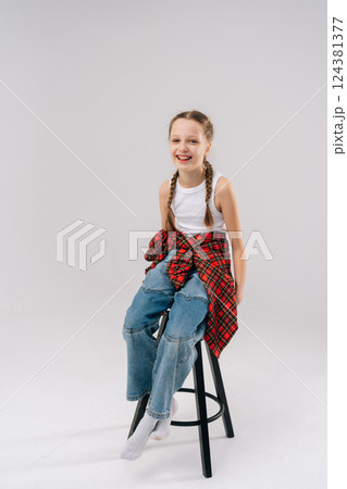 Cheerful preteen girl with long braids, wearing white tank top, red checkered shirt and jeans, sitting on black stool against white background, radiating confidence, smiling looking at camera. 124381377