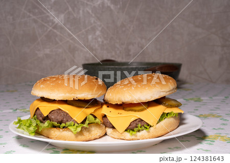 Two cheeseburgers with sesame buns, beef patties, melted cheddar, lettuce, pickles, and tomato sit on a white plate, ready for serving. Two cheeseburgers with sesame buns, beef patties, melted cheddar, lettuce, pickles, and tomato sit on a white plate, ready for serving. 124381643