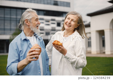 Caucasian elderly couple standing on a grass in summer. Man and woman holding a two croissants in hands. Woman wearing white shirt and man blue one. Caucasian elderly couple standing on a grass in summer. Man and woman holding a two croissants in hands. Woman wearing white shirt and man blue one. 124384239