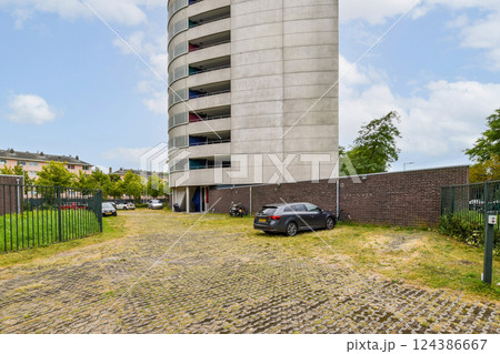 A sleek modern residential building towers over a structured parking area surrounded by greenery and a boundary wall, highlighting urban architecture. 124386667