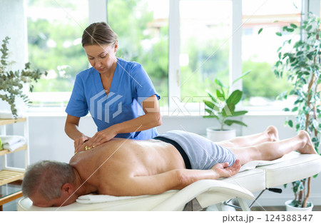 Skilled middle-aged masseuse conducting back massage for man client Skilled middle-aged masseuse conducting back massage for man client 124388347