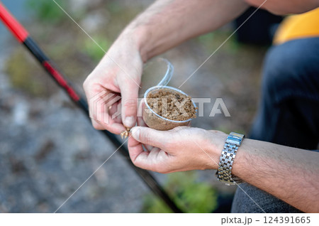 Angler is preparing groundbait for carp fishing, holding a plastic container with the bait and a hook in his hands 124391665