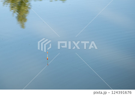 Fishing float with orange tip waits patiently on a calm blue water surface, reflecting trees and sky 124391676