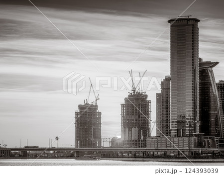 Monochrome city skyline with tall skyscrapers reflecting on calm water under a cloudy sky, Dubai Marina bay UAE 124393039