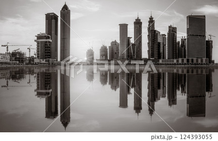 Monochrome city skyline with tall skyscrapers reflecting on calm water under a cloudy sky, Dubai Marina bay UAE 124393055